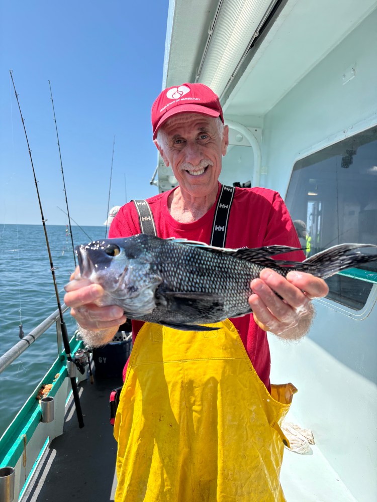 Man in red cap and yellow overalls holding a fish on a boat.