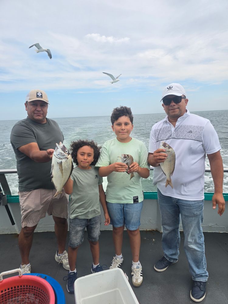 Three men and a boy holding fish on a boat, with seagulls and ocean in the background.