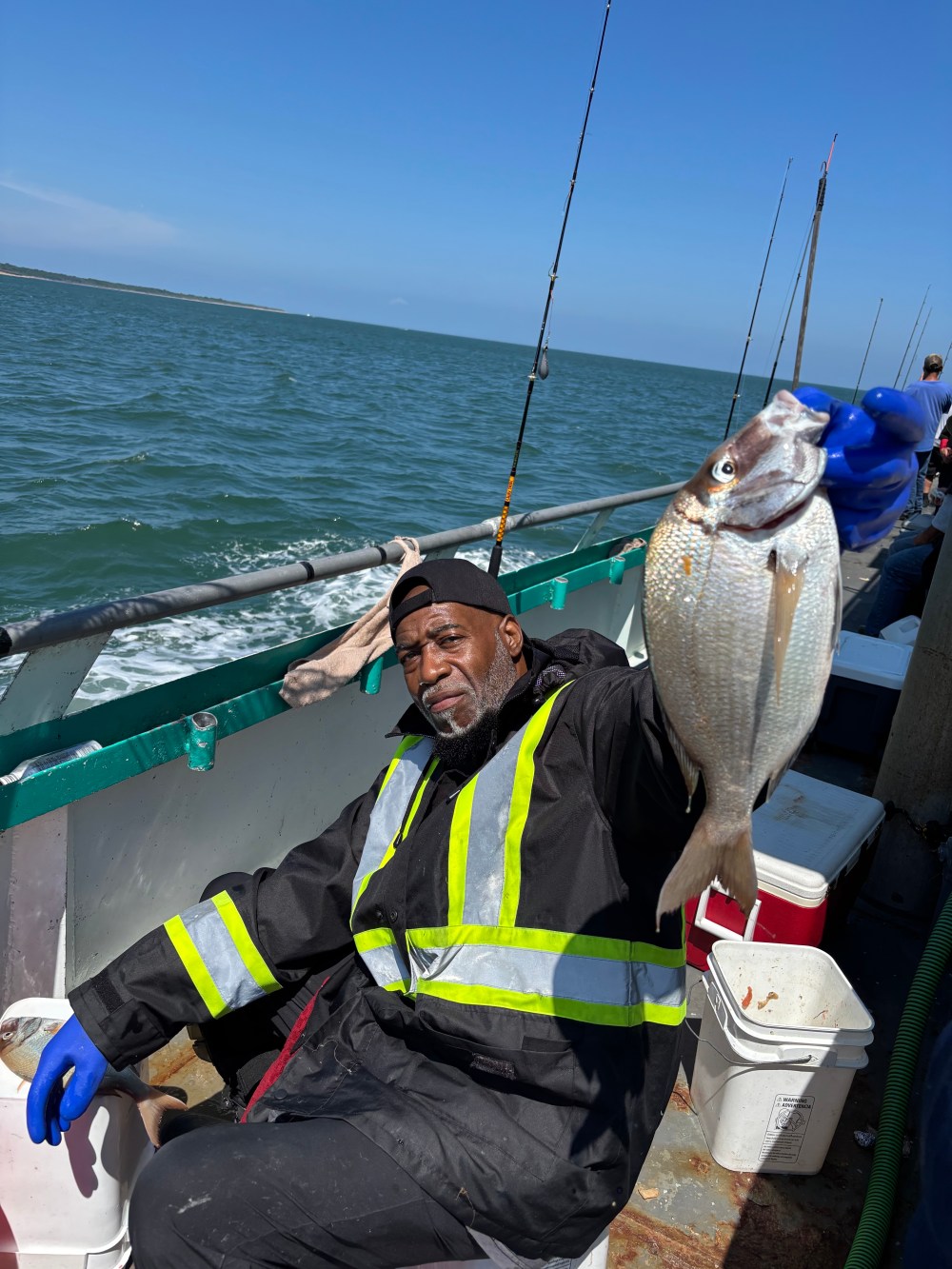 Person on a boat holding a fish, wearing a black jacket and blue gloves.