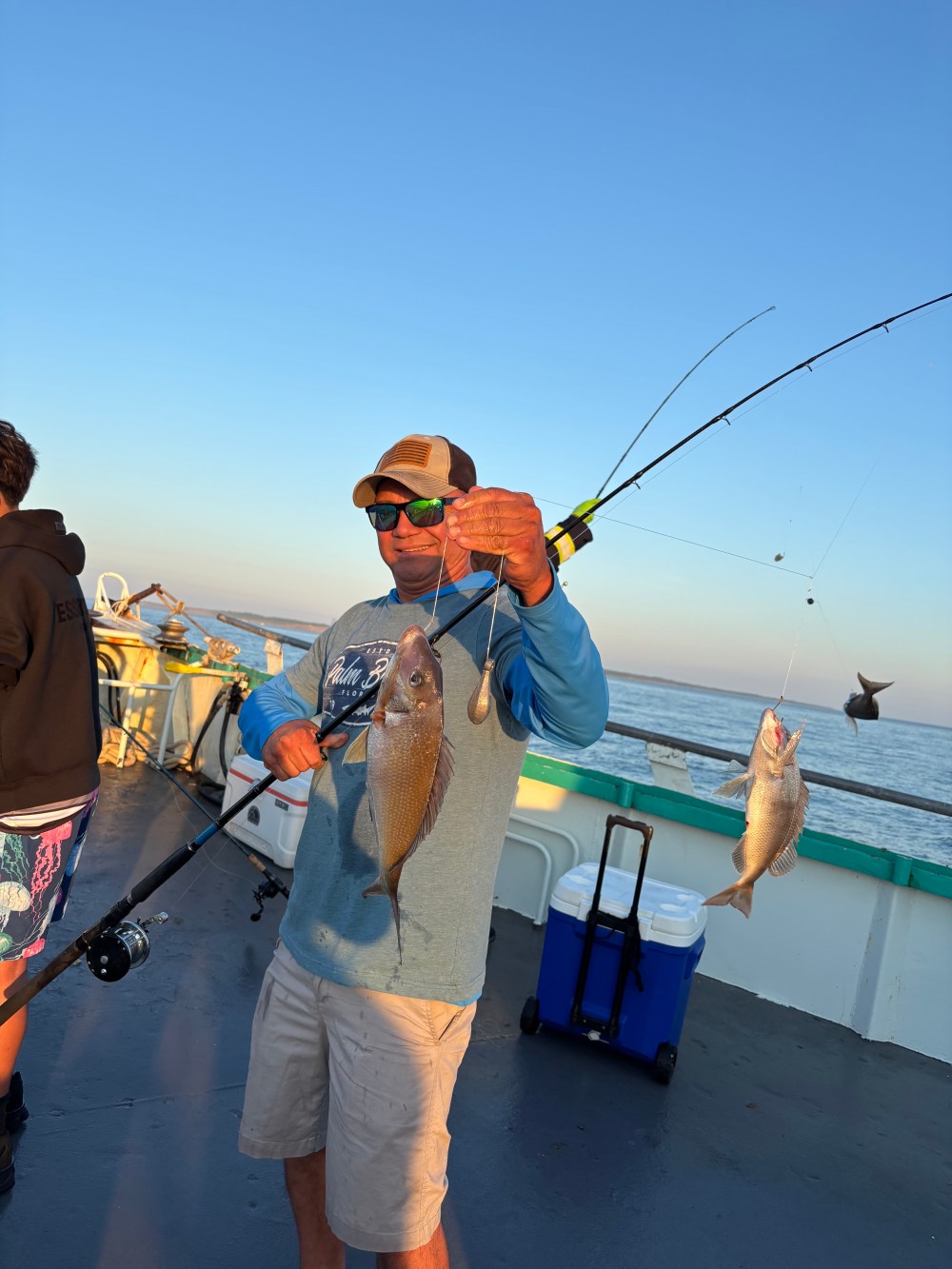 Man holding two fish on a boat under a clear sky.