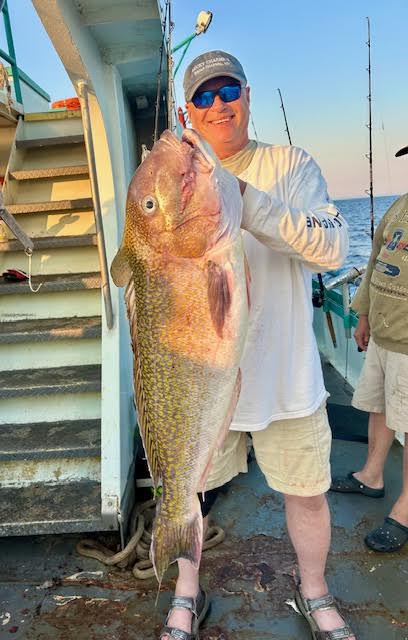 Man holding a large fish on a boat with fishing rods in the background.
