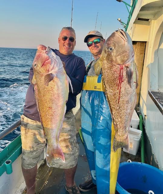 Two men on a boat holding large fish, smiling at the camera.