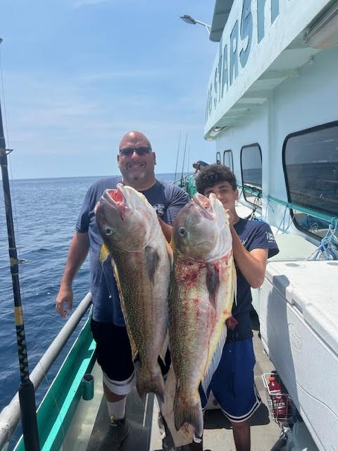Two people holding large fish on a boat deck with ocean in background.