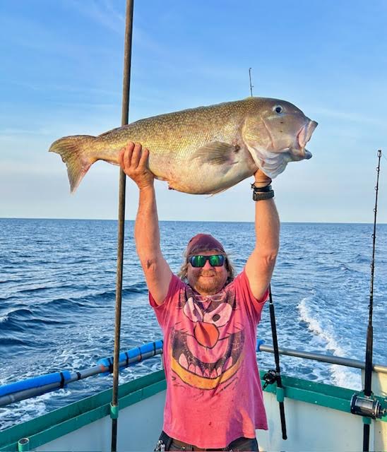 Person on a boat holding a large fish overhead with the ocean in the background.