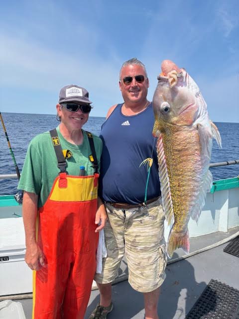 Two men on a boat hold a large fish against a blue sky backdrop.