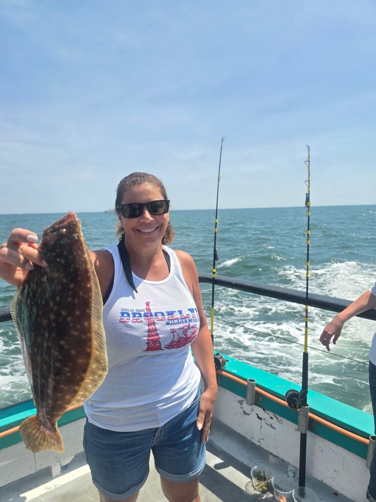 Woman on a boat holds a flounder, smiling, with fishing rods and ocean in the background.