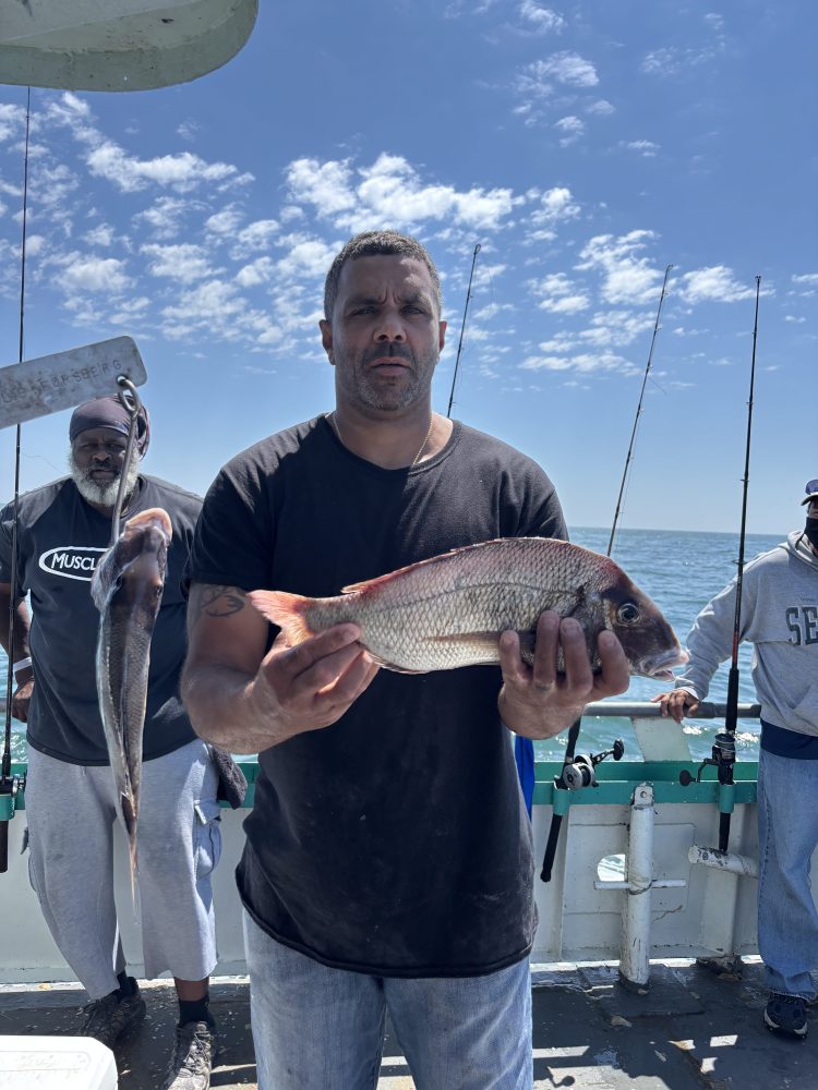 Man holding a large fish on a boat with others and fishing rods in the background.