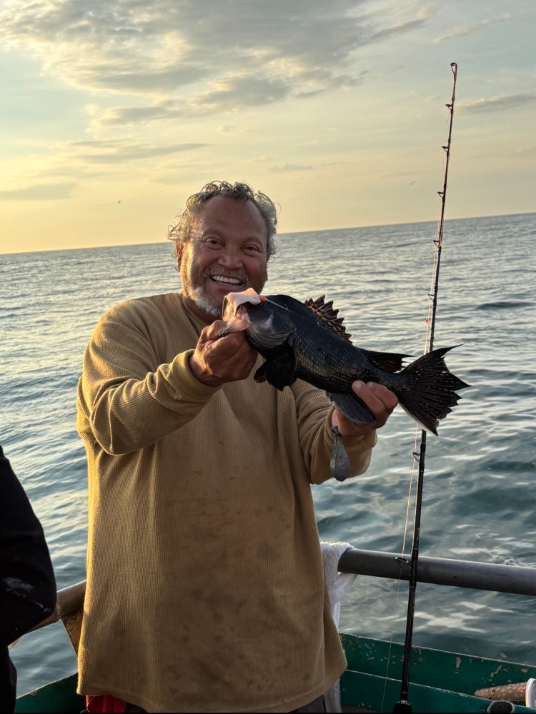 Smiling man holding a fish on a boat at sunset with a fishing rod nearby.