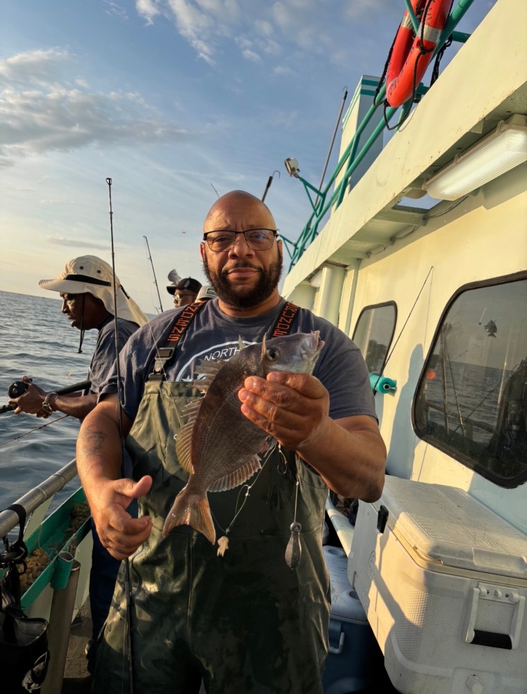 Person holding a fish on a boat, with another person fishing in the background.