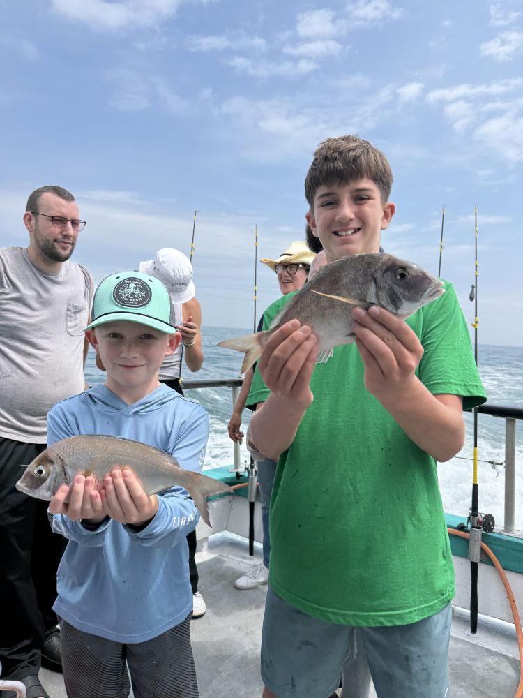 Two children proudly holding fish on a boat, smiling with fishing rods in the background.