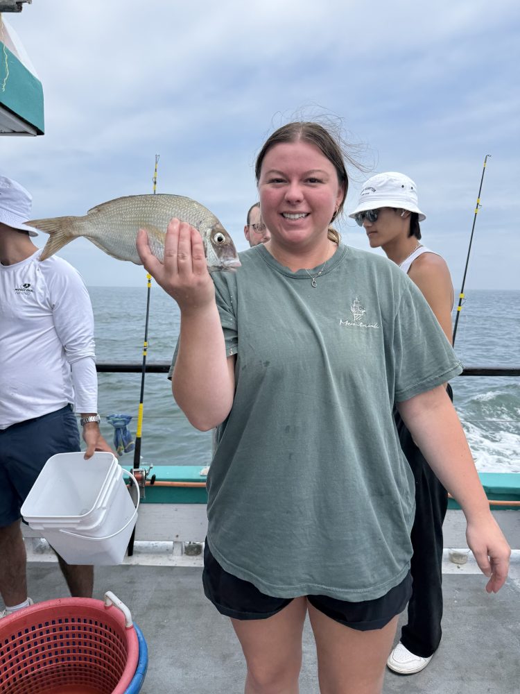 Smiling person holds a fish on a boat with ocean in the background.