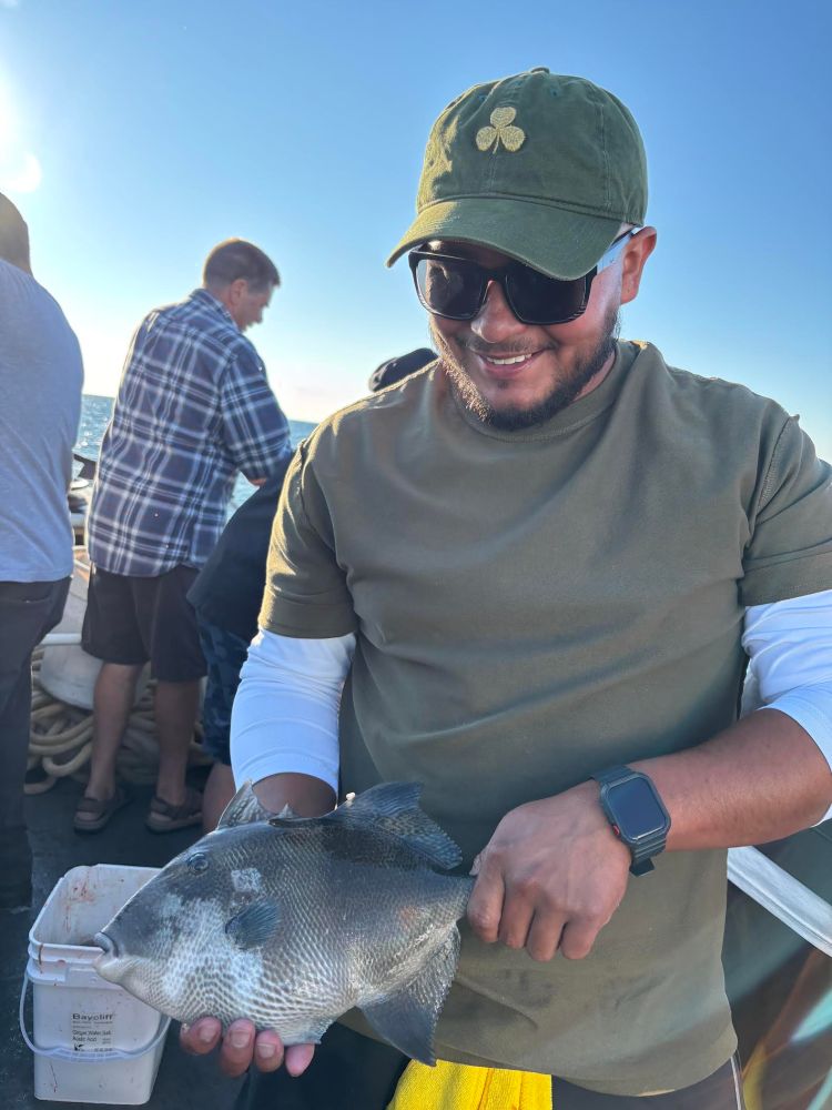 Man in cap holding a fish on a boat, with ocean in background.