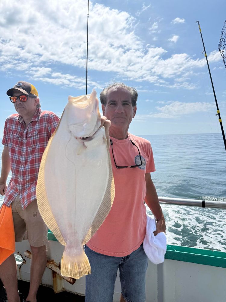 Man holding a large fish on a boat with another person and ocean in the background.