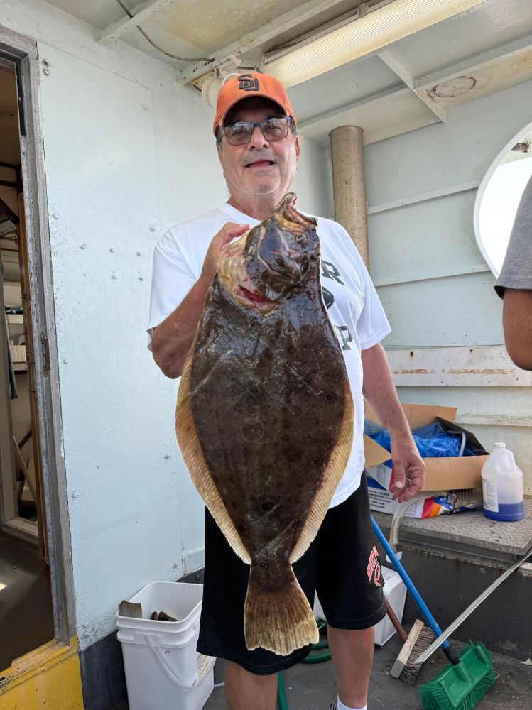 Man holding a large flatfish on a boat, wearing sunglasses and a cap.