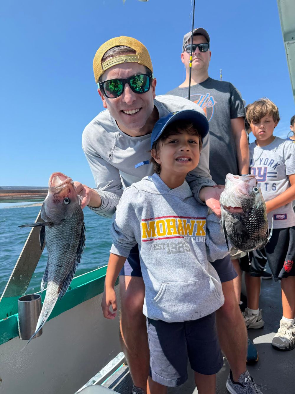 Man and boy on a boat holding two large fish, smiling with others in the background.