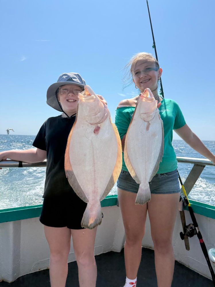 Two people on a boat holding large flatfish, smiling under clear blue sky.
