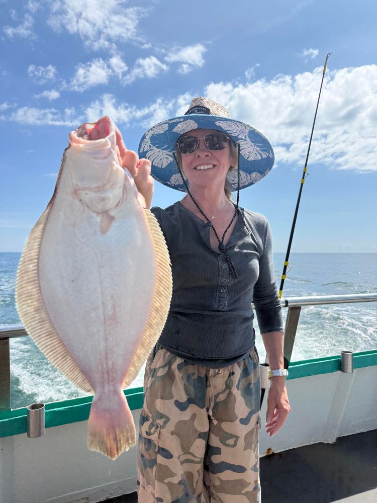 Person on a boat holding a flat fish, wearing a wide-brimmed hat and sunglasses, with ocean in the background.