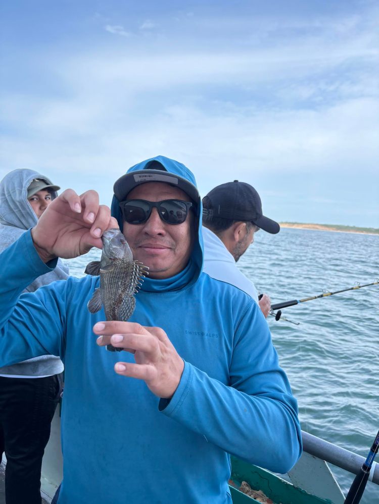 Man in blue hoodie holding a small fish on a boat with others fishing nearby.