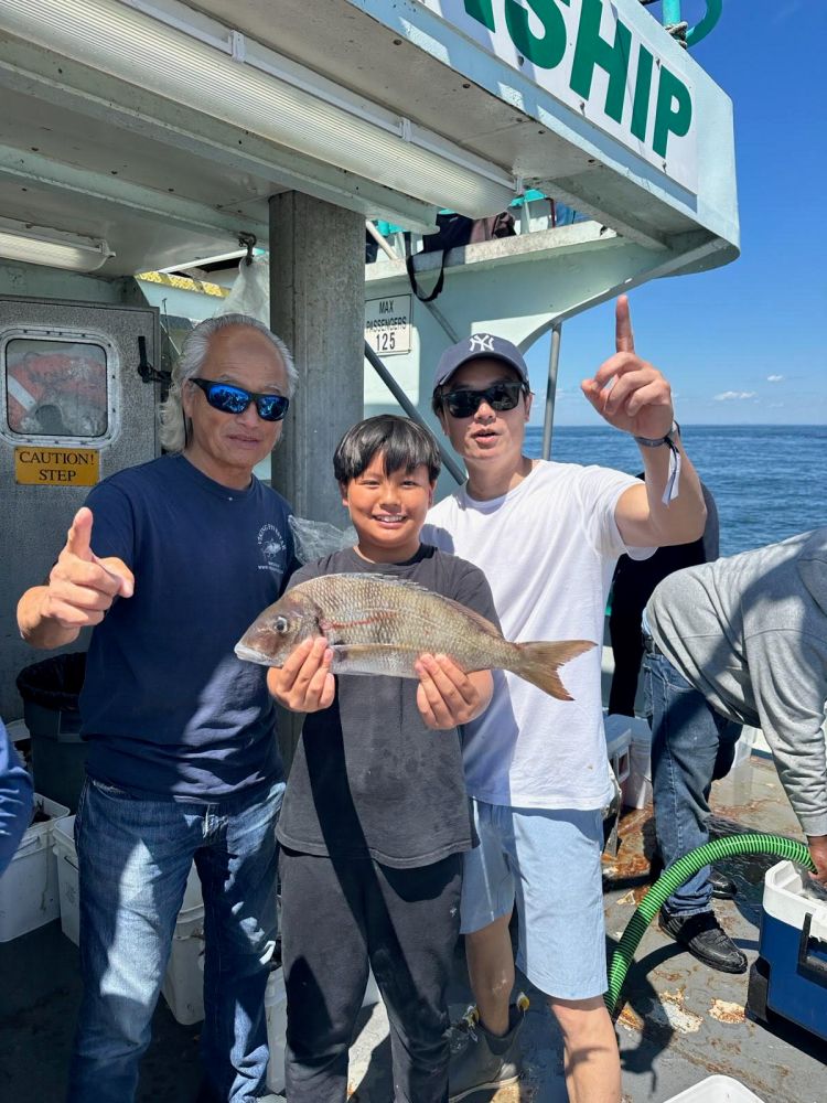 Three people on a boat, smiling and holding a fish, with ocean in the background.