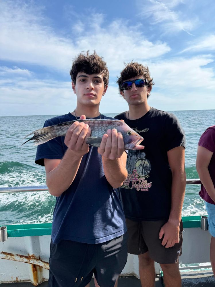 Two people on a boat, one holding a fish, with ocean and sky in the background.