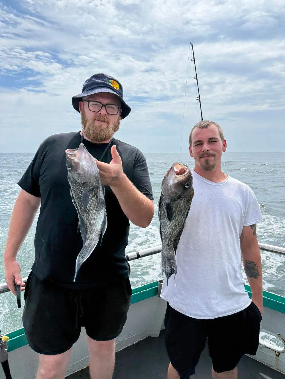 Two men on a boat holding fish, with ocean in background.