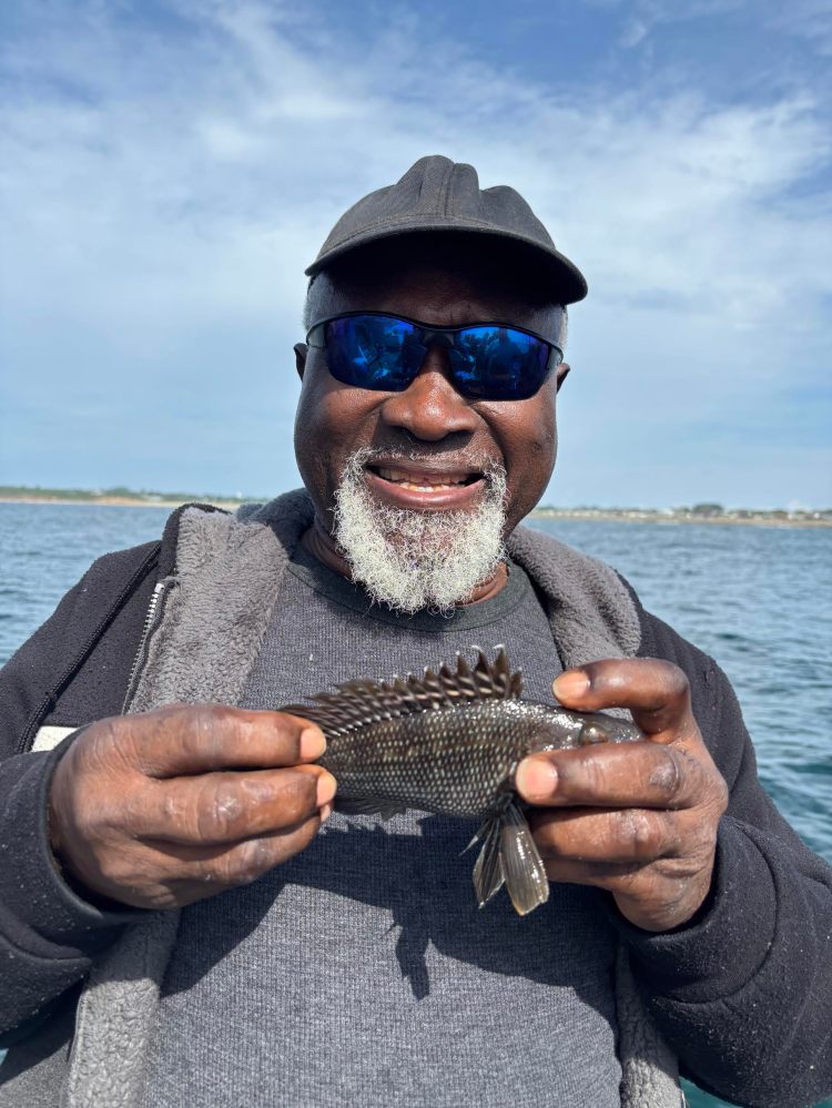 Man smiling, holding a small fish by the water, wearing sunglasses and a cap.