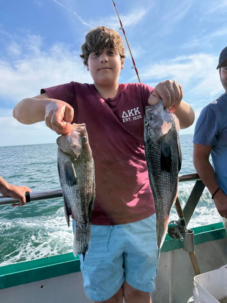 Person on a boat holding two fish, ocean in the background, clear sky.