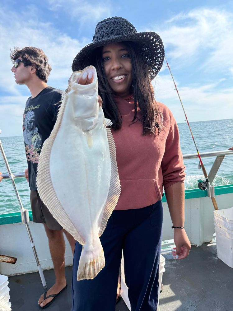 Person holding a flat fish on a boat with another person nearby and ocean in the background.