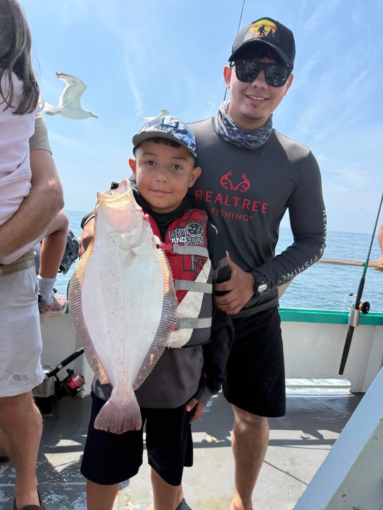 Child holding a large fish on a boat, with an adult and seagulls in the background.