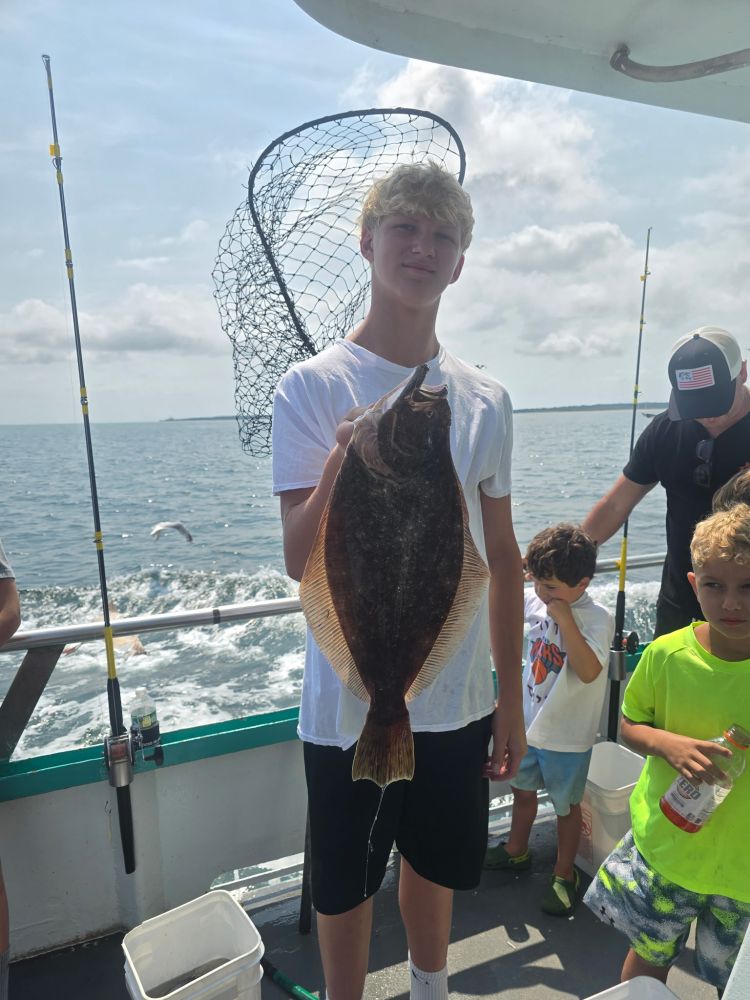 Young person holding a large fish on a boat, with water and fishing poles in the background.