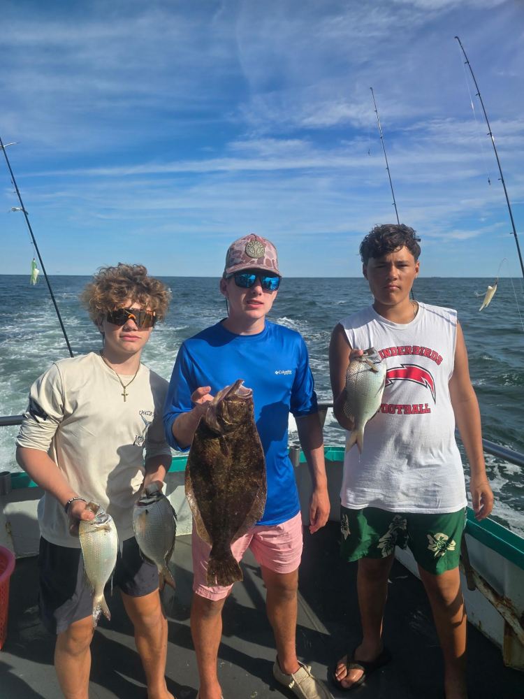 Three people on a boat holding fish with blue sky and ocean in the background.