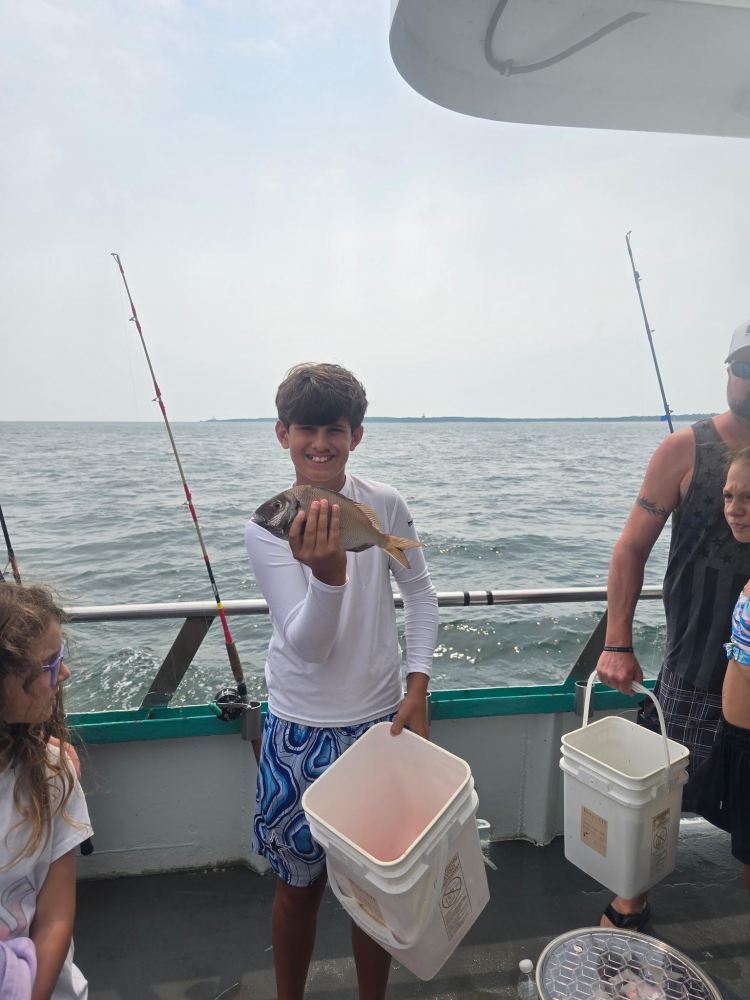Boy on a boat holding a fish, surrounded by people and fishing rods.