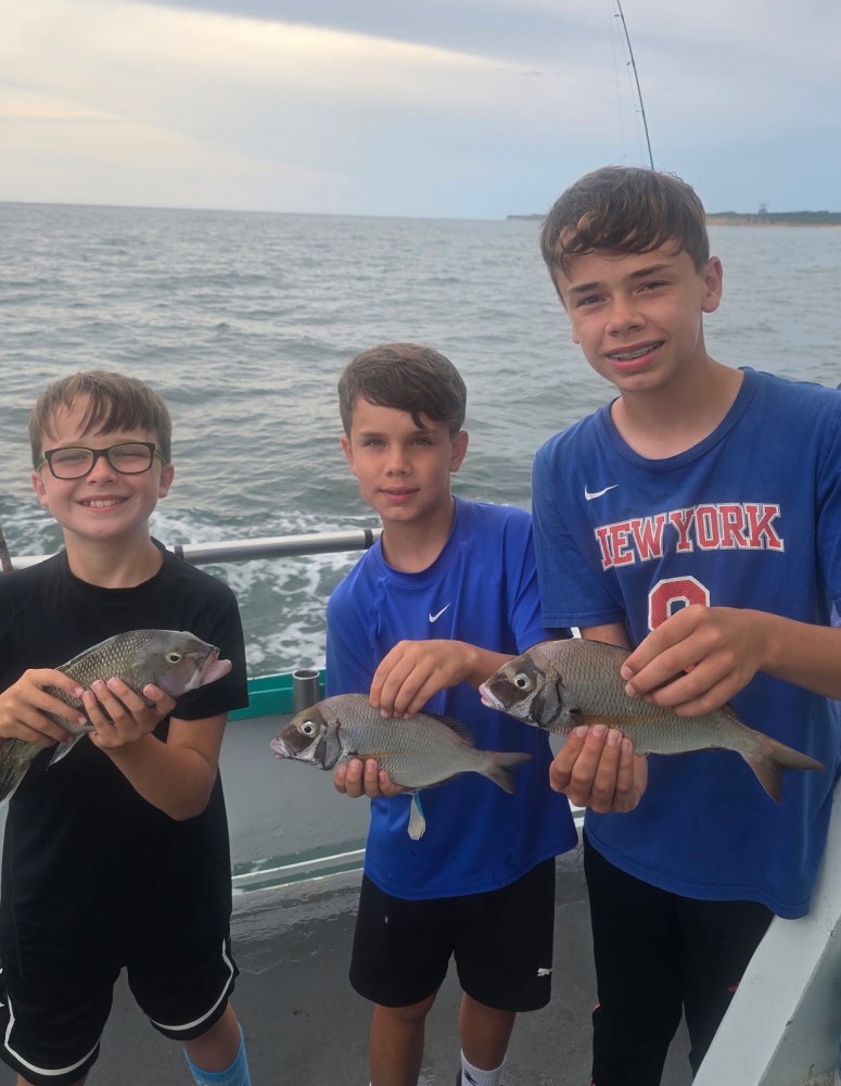 Three boys on a boat holding fish, overcast sky and ocean in background.