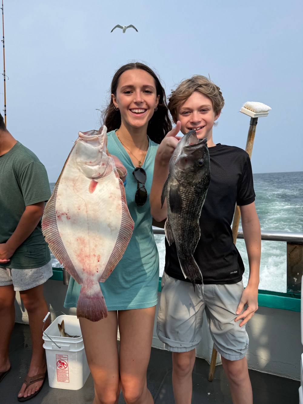 Two people holding large fish on a boat with a seagull overhead.