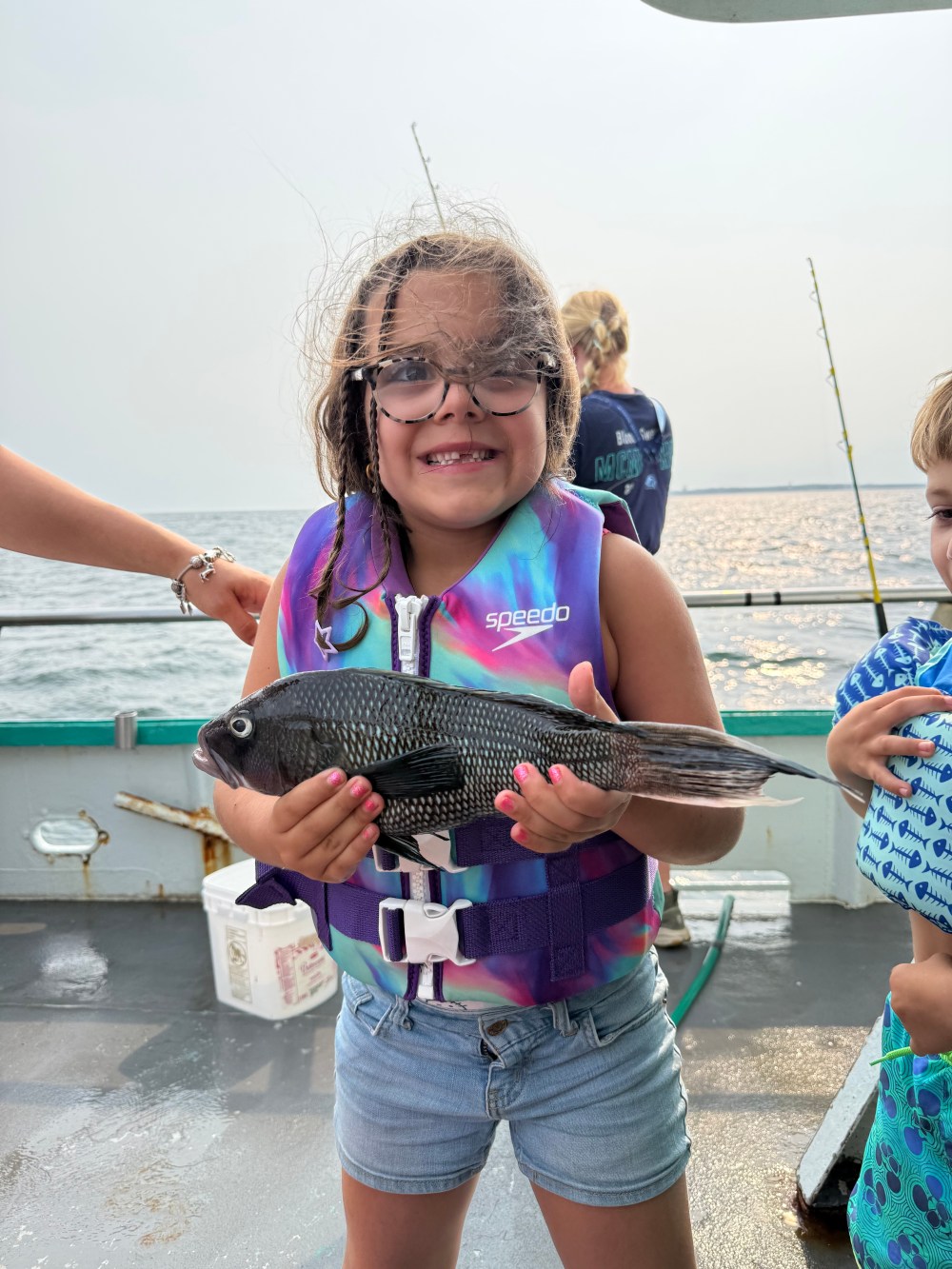Girl in life vest holding a fish on a boat, smiles with two people nearby.