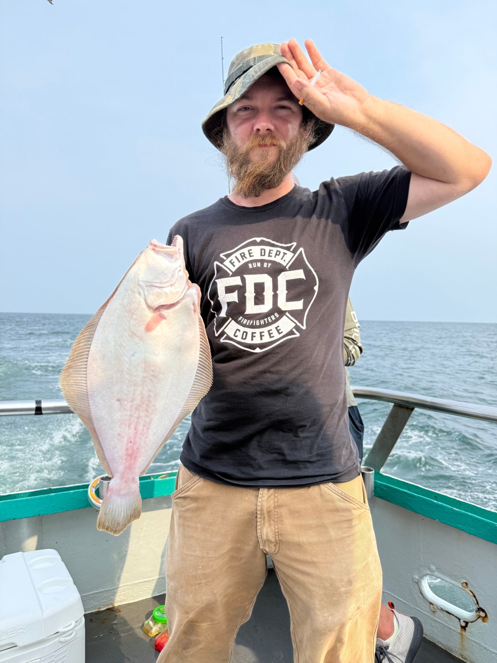 Person on boat holding a flat fish and saluting, wearing a hat and dark shirt.