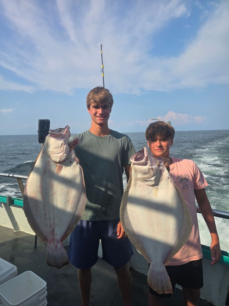 Two people holding large fish on a boat with ocean in the background.
