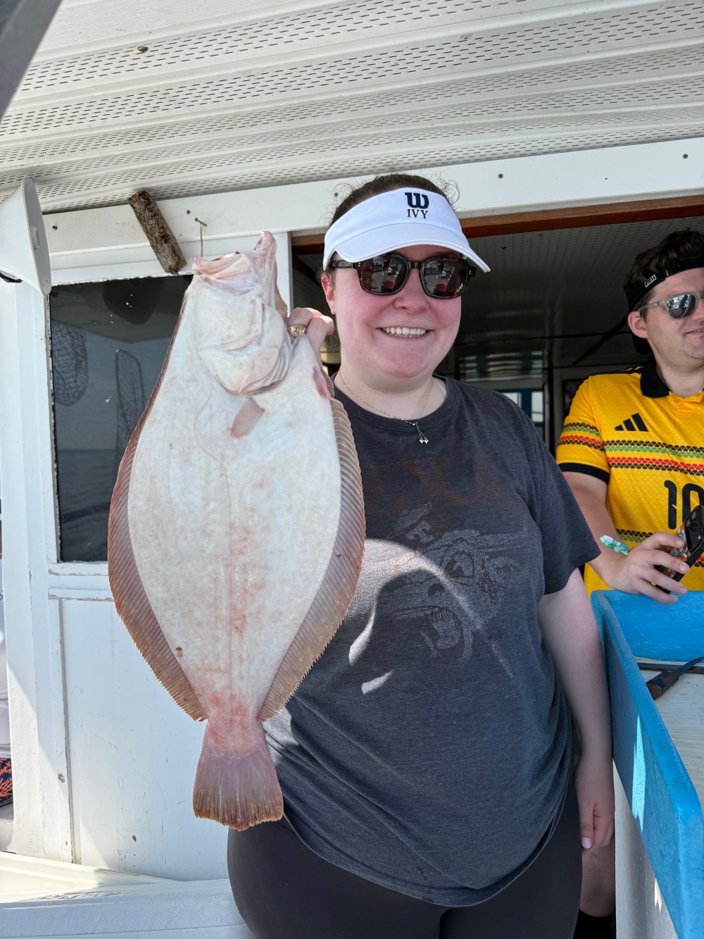 Person holding a large flat fish on a boat, smiling, wearing sunglasses and a visor.