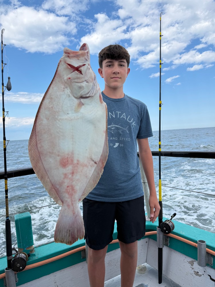 Boy holding a large fish on a boat with fishing rods and ocean in the background.