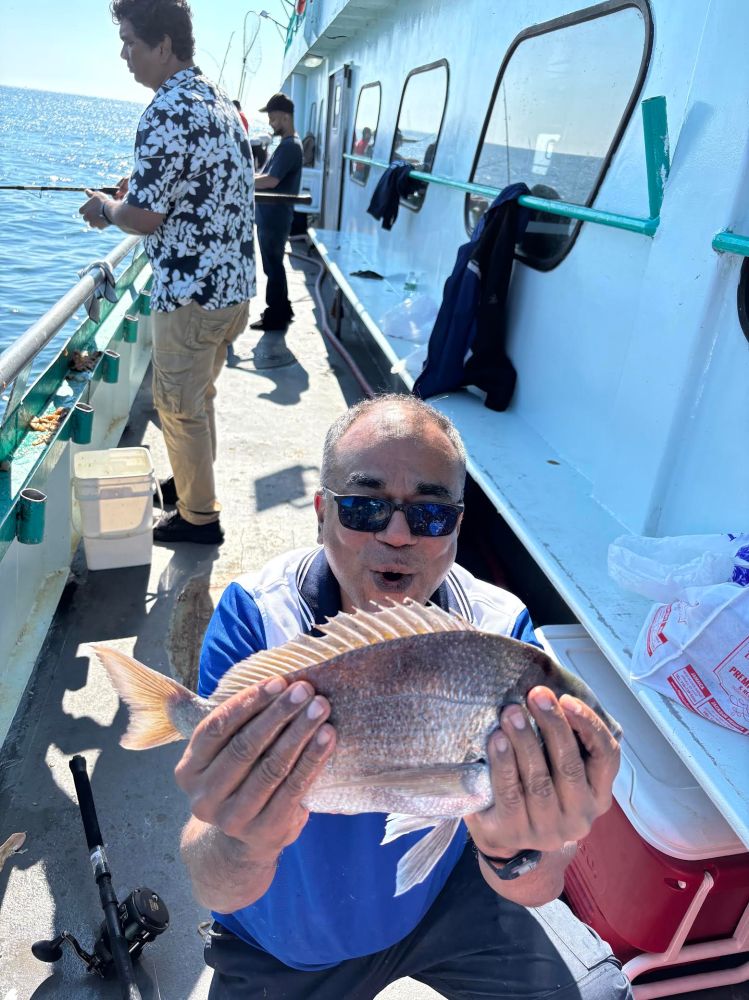 Man on boat holds a fish, another person fishes in the background.