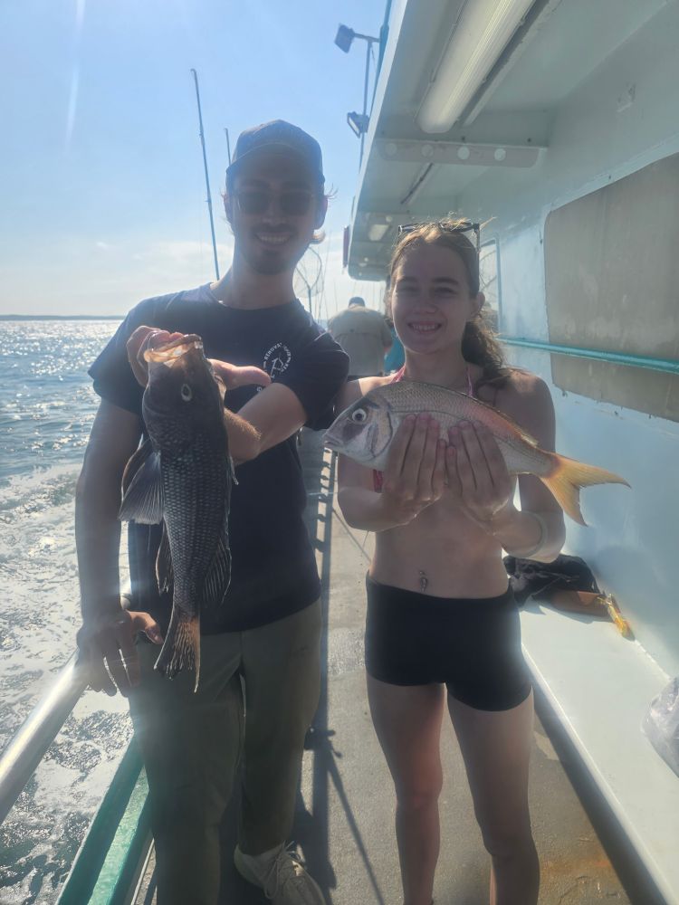 Two people on a boat holding fish, with ocean in the background.