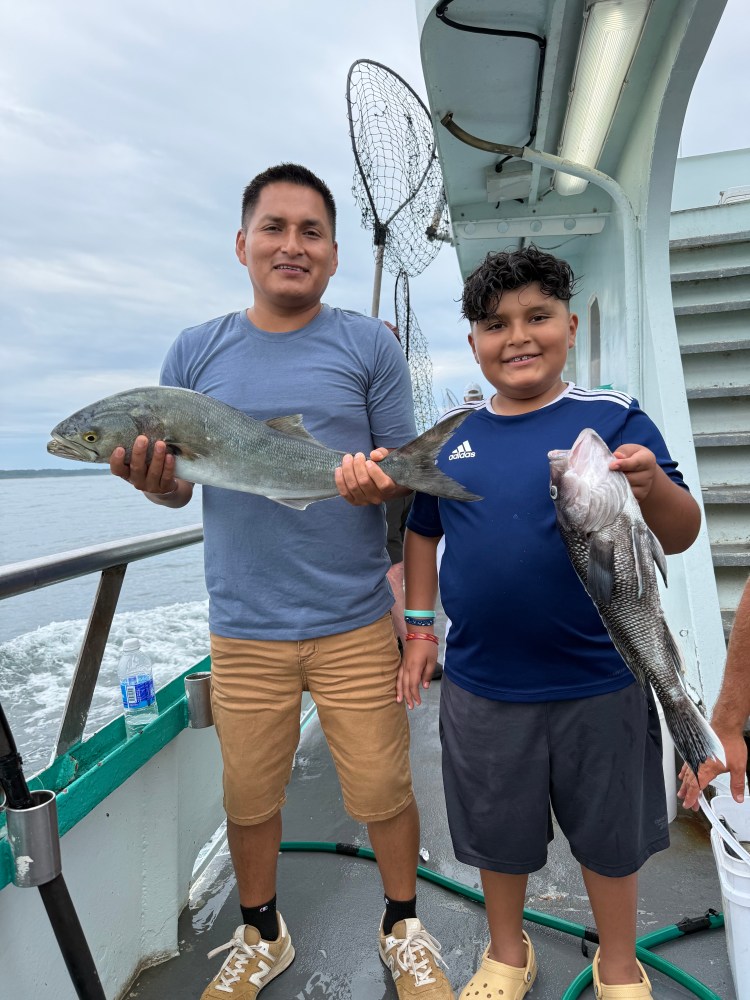 Two people on a boat holding fish, one wearing a blue shirt and shorts, the other in a striped shirt and sandals.