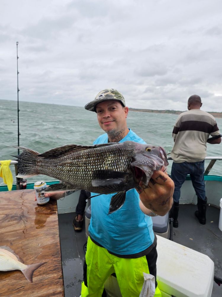 Person on a boat holding a large fish, with the sea and another person visible in the background.