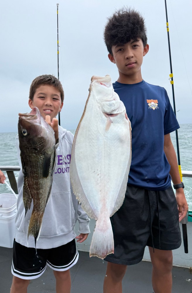 Two boys on a boat holding a large fish each, with fishing rods in the background.