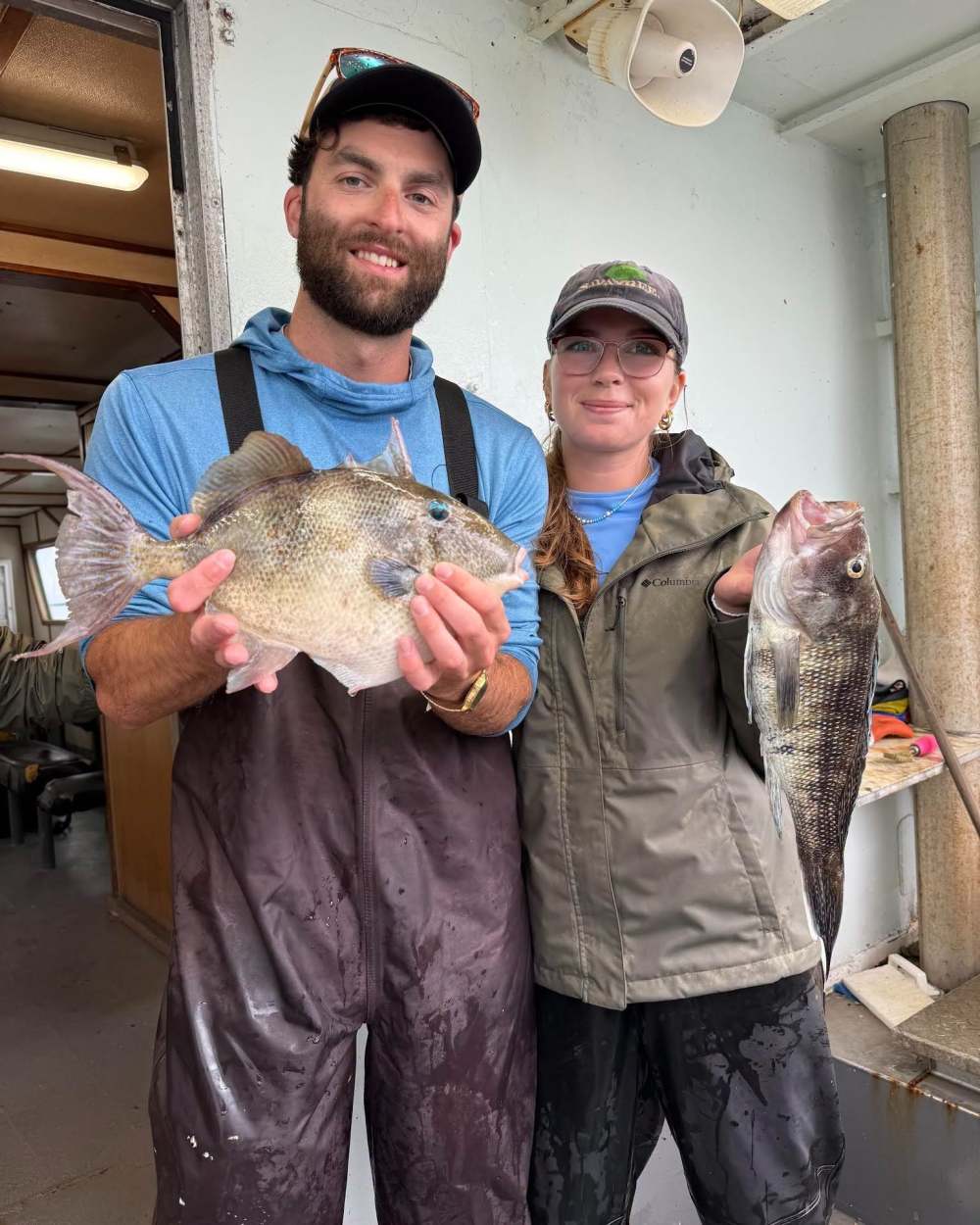 Two people on a boat holding caught fish, both smiling.