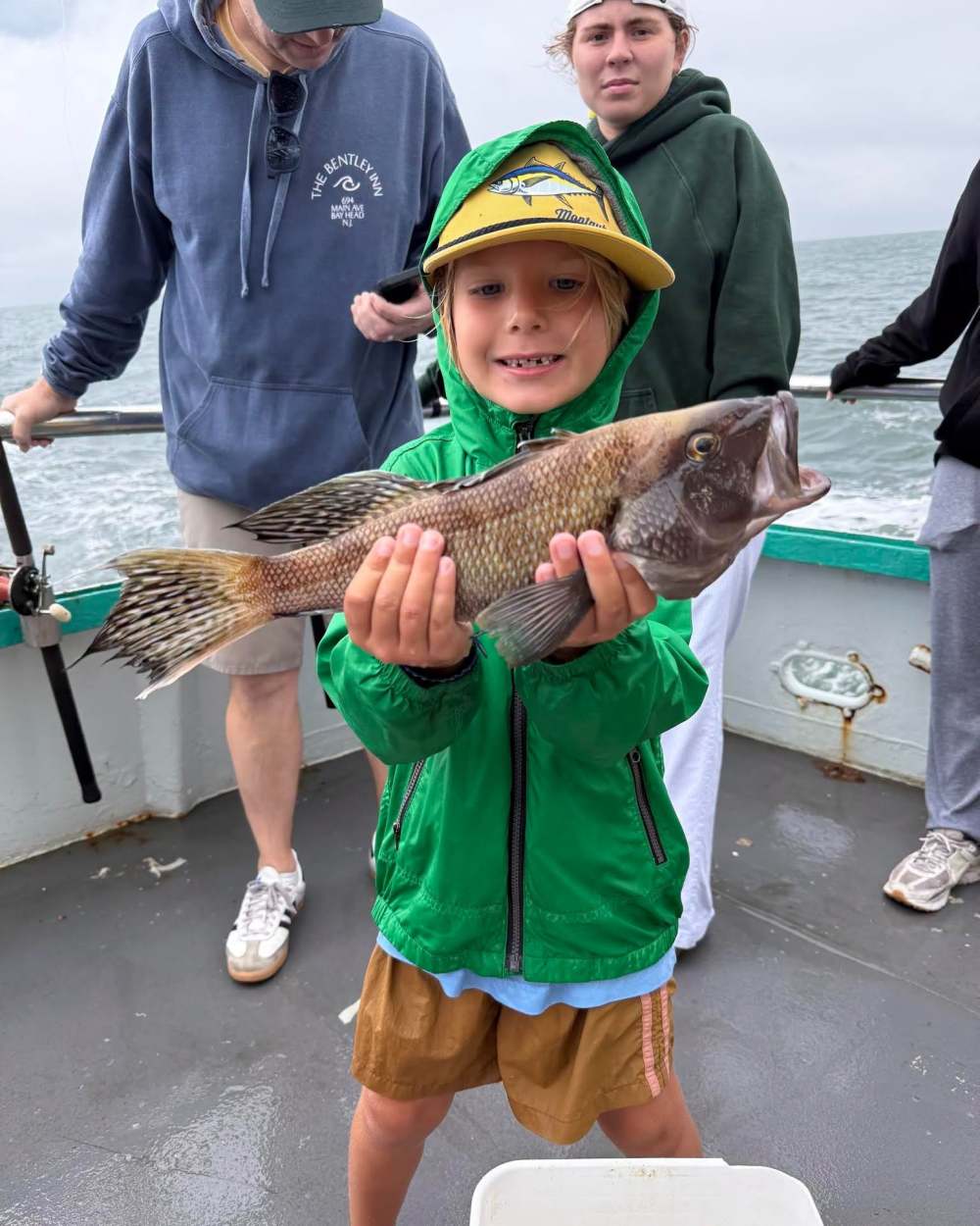 Child in a green jacket proudly holds a fish on a boat, with a cloudy sea background.
