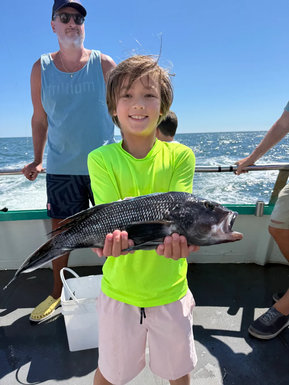 Child in neon shirt holding a large fish on a boat with ocean background.
