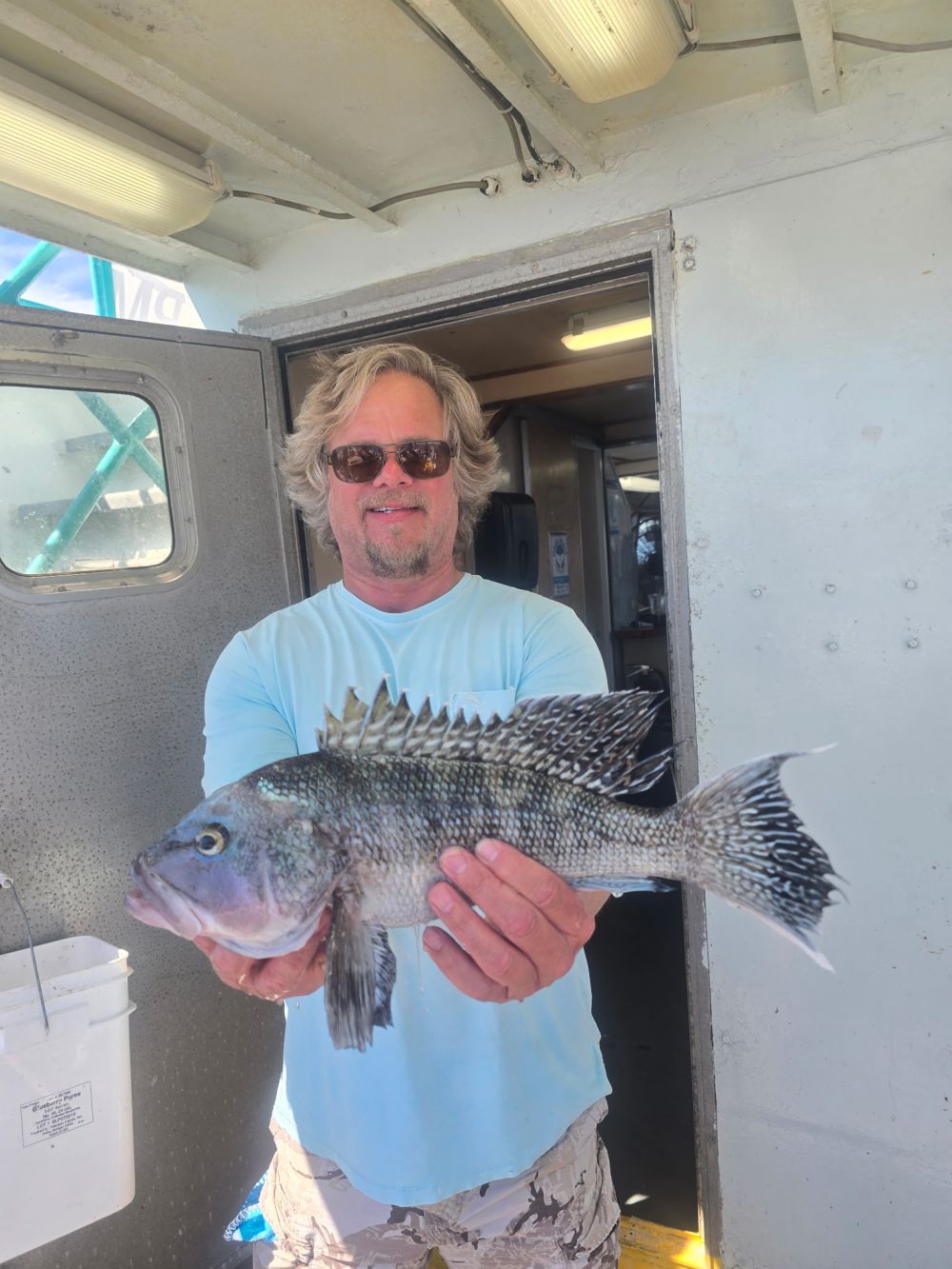 Man holding a large fish on a boat, wearing sunglasses and a light blue shirt.