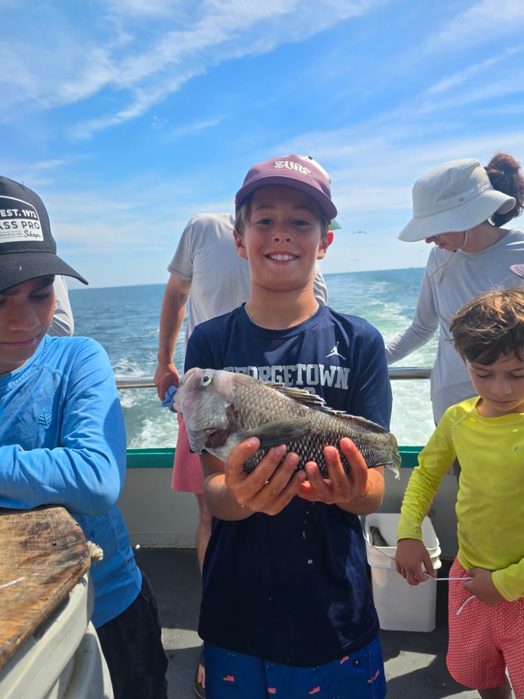 Boy on a boat holding a fish, smiling, with others around and ocean in the background.