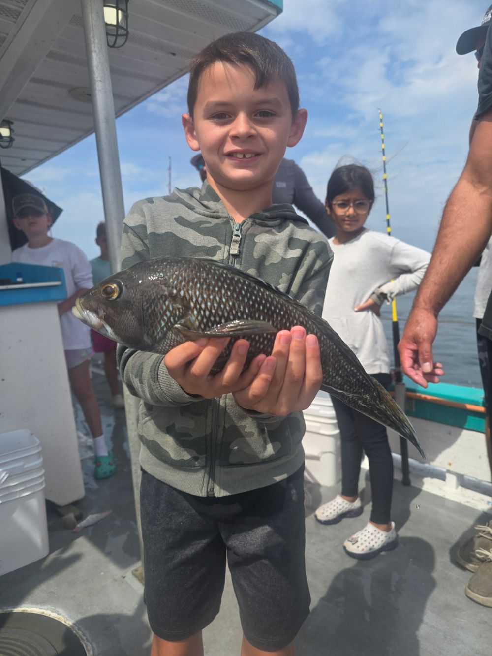 Child on a boat holding a fish, with people and fishing rods in the background.